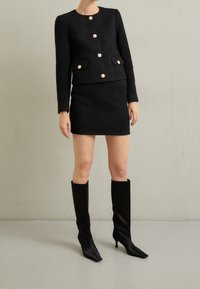 Zwarte textuurjacket met zes goudkleurige knopen en twee voorzakken, gecombineerd met een bijpassende zwarte mini-rok en zwarte kniehoge laarzen.