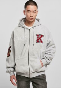 Sweat à capuche gris zippé avec une grande lettre "K" rouge brodée et un patch graphique sur la manche gauche. Capuche avec cordon de serrage, poches avant.