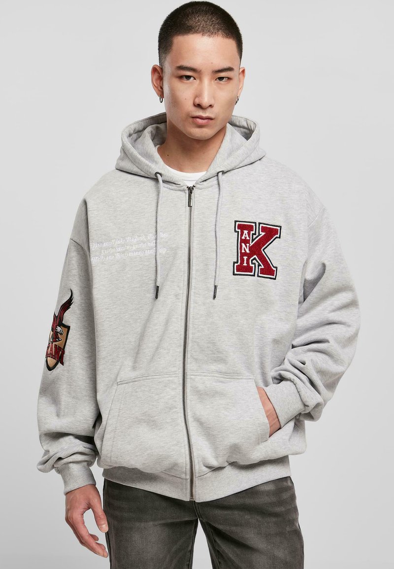 Sweat à capuche gris zippé avec une grande lettre "K" rouge brodée et un patch graphique sur la manche gauche. Capuche avec cordon de serrage, poches avant.