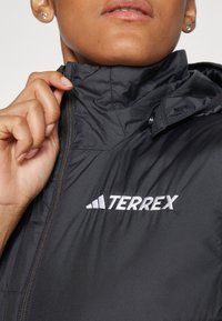 Svart pufferjacka med hög krage, har dragkedja och vit "TERREX"-logotyp. Slät, glänsande tyg med en lätt design.