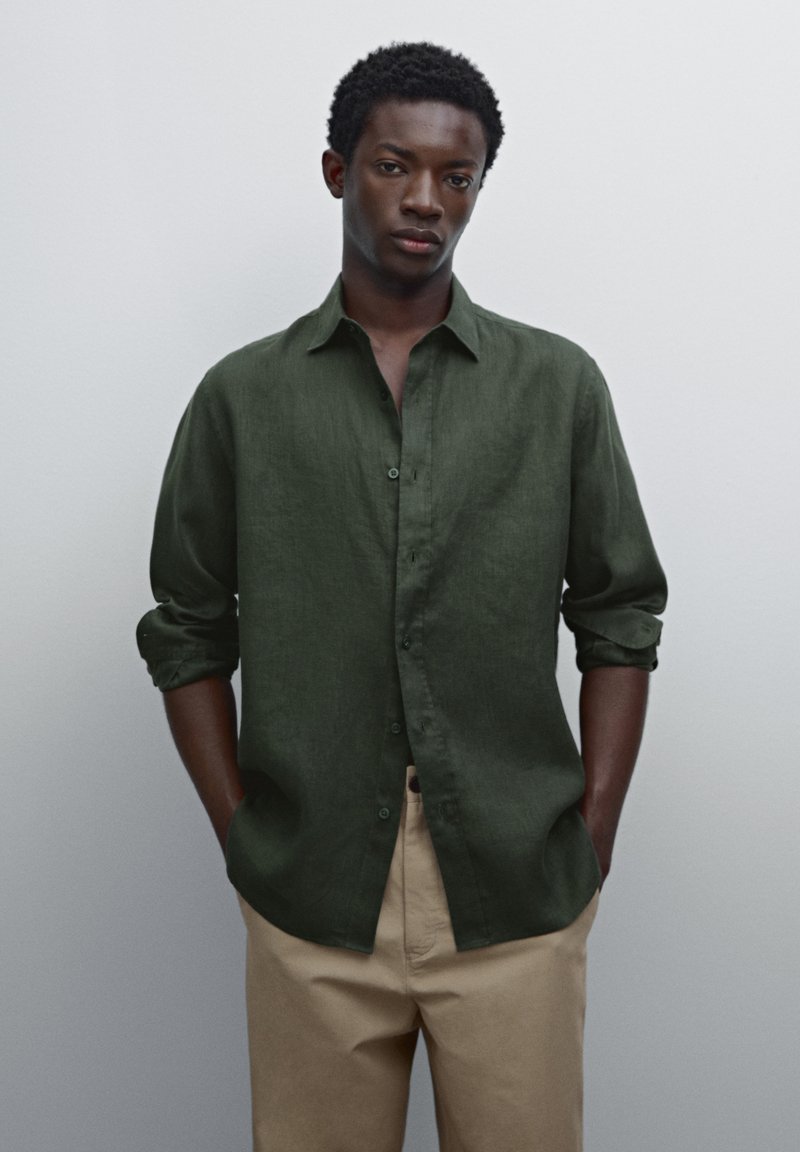 Chemise en lin vert foncé avec un devant à boutons, manches retroussées et coupe décontractée, associée à un pantalon beige clair. Texture lisse.