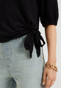 Czarna, teksturowana bluzka z bufkami, związana w talii, w połączeniu z jasnoniebieskimi dżinsami i złotą bransoletką na nadgarstku.