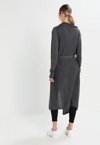 Cardigan largo gris de tela suave con mangas largas, cintura ceñida y un dobladillo a media longitud. Llevado sobre leggings negros y zapatillas blancas.