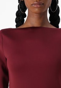 Robe ajustée bordeaux avec un décolleté bateau et des manches longues, fabriquée en tissu lisse. Présente de grandes boucles d'oreilles créoles en arrière-plan.