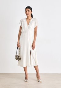 Weißes Midi-Kleid mit tiefem V-Ausschnitt, kurzen Ärmeln und seitlichem Schlitz. Kombiniert mit einer Handtasche im Leopardenmuster und metallischen High Heels.