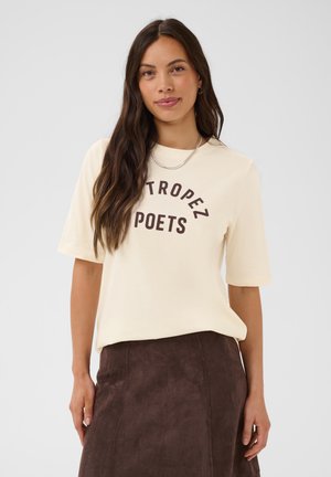 Žena s dlhými hnedými vlasmi má na sebe krémovú košeľu s nápisom "ST TROPEZ POETS" a hnedú sukňu, stojí pred jednoducho svetlým pozadím.