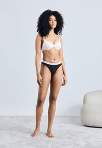 Reggiseno imbottito bianco e slip bikini neri con fascia in vita con logo. La modella è in piedi su una superficie chiara e testurizzata, con le braccia rilassate lungo i fianchi.