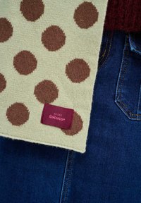 Pletená šatka v krémovej farbe s hnedými bodkami. Obsahuje bordovú štítok s nápisom "Naoko Knitwear." Pozadie tvorí tmavomodrý denim.