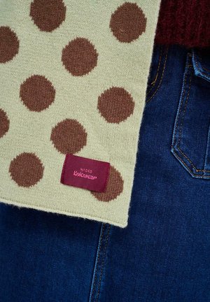 Dziany szalik w kremowym kolorze z brązowymi kropkami. Zawiera bordową metkę z napisem "Naoko Knitwear." Tło obejmuje ciemnoniebieski denim.