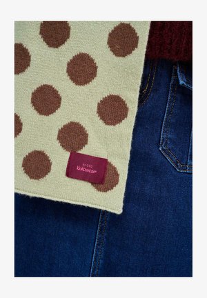 Dziany szalik w kremowym kolorze z brązowymi kropkami. Zawiera bordową metkę z napisem "Naoko Knitwear." Tło obejmuje ciemnoniebieski denim.