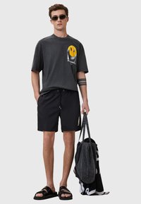 T-shirt di cotone nera con una grafica di un sorriso giallo e il testo "ALLSAINTS", abbinata a pantaloni neri e sandali neri, con una borsa a tracolla nera.