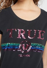 Black T-shirt with shiny pink sequined text "TRUE" above a multicolored sequined stripe and a circular emblem with an upward arrow.