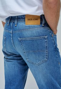 Vaqueros de mezclilla azul con un lavado claro, que presentan una etiqueta de cuero con la inscripción "SALSA JEANS". Dos bolsillos traseros con detalles de costura curva.