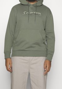 Felpa con cappuccio verde oliva con tasca a canguro frontale, polsini e fondo a coste, con logo Calvin Klein in bianco. Realizzata in tessuto morbido.