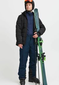 Chaqueta de plumas negra, pantalones aislantes azul marino, capa base azul. Sostiene esquís verdes y bastones. Botas de esquí negras y casco con visera naranja.