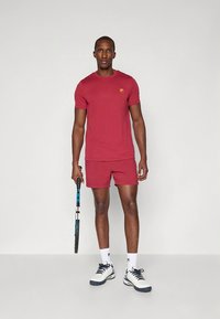 Conjunto de camiseta de manga corta y pantalones cortos color granate, hecho de algodón, con un pequeño logotipo. El modelo sostiene una raqueta de tenis y lleva zapatillas blancas.