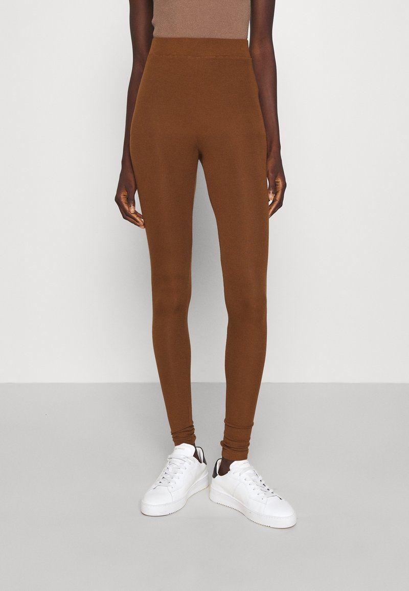 Leggings marron taille haute, ajustés, portés avec des baskets blanches et un top marron sans manches sur un fond gris clair uni.