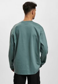 Long-sleeve shirt in muted teal, made from soft fabric. Features a crew neck, relaxed fit, and slightly curved hemline.
