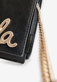 Juodos odos rankinė su auksiniu metaliniu "la" akcentu. Turi grandinėlės dirželį ir mažą žymeklį su užrašu „ROCKETT GEORGE“ auksiniais rašmenimis.