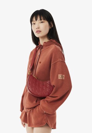Red leather handbag with diamond pattern, curved shape, and gold-tone hardware, paired with a rust-colored hoodie and shorts.