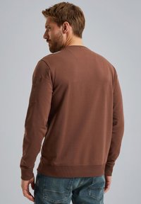 Sweatshirt marron à manches longues, décolleté rond et poignets côtelés. Il présente une poche latérale sur la manche gauche. Porté avec un jeans en denim bleu.