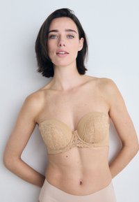 Strapless nude bh med blonde detaljer, glatt stoff og en dekorativ gullaccent i midten. Klassisk fasong med en myk tekstur.