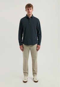 Donkerblauw overhemd met een gemêleerd uiterlijk, gedragen met lichtbeige broek en witte sneakers. Losstaande figuur op een neutrale achtergrond.