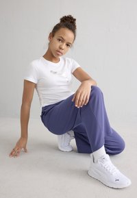 Valge lühikeste varrukatega t-särk hõbedase logoga, koos tekstuursete lillade pükstega ja valgete tossudega, istumisasendis heledal pinnal.