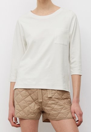 Nainen, joka käyttää valkoista kolmivartista paitaa, jossa on rintatasku, ja beigejä tikattuja shortseja, joissa on kiristysnauhat.