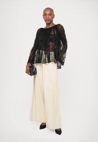 Schwarzes Spitzenoberteil mit langen Ärmeln, transparentem Stoff und Falten-Design, kombiniert mit hochtaillierten, cremefarbenen, gefalteten Hosen und schwarzen spitzen Pumps.