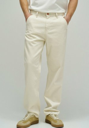 Hombre con pantalones rectos color crema y zapatillas beige con cordones blancos, manos en los bolsillos, de pie contra un fondo liso.