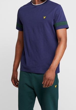 Homme portant un t-shirt bleu marine avec des rayures vertes sur les manches et un pantalon vert foncé, tous deux ornés d'un petit logo d'oiseau jaune.