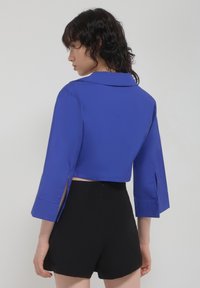 Camicia blu tagliata con colletto e maniche ampie, con fessure laterali. Abbinata a pantaloni shorts neri. Texture liscia, design aderente.