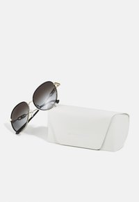 Occhiali da sole neri e oro con lenti grigie sfumate, forma ovale e accessori decorativi. Accompagnati da una custodia bianca testurizzata.