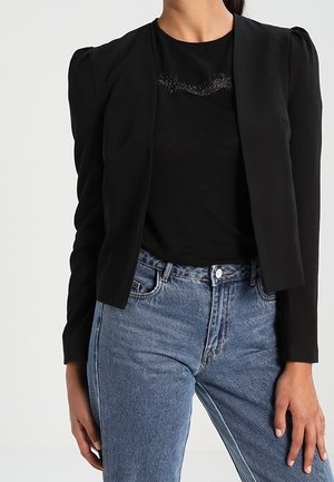 Kvinde iført en figursyet sort blazer over en sort top med blonde detaljer, kombineret med højtaljede blå jeans mod en ensfarvet baggrund.