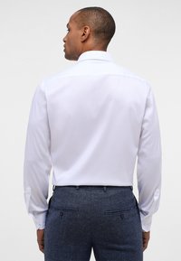 Camicia bianca a maniche lunghe con colletto classico, tessuto morbido e polsini abbottonati, abbinata a pantaloni strutturati di colore blu scuro.