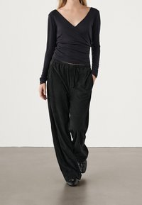 Czarna bluzka z długim rękawem i głębokim dekoltem w kształcie litery V, zestawiona z luźnymi czarnymi spodniami. Oba elementy wykonane z miękkiego, gładkiego materiału.