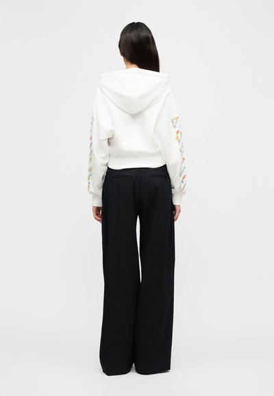 Λευκό φούτερ με κουκούλα και κορδόνι, με πολύχρωμα γραφικά στα μανίκια, σε συνδυασμό με μαύρο παντελόνι φαρδύ. Ομαλή υφή υφάσματος.