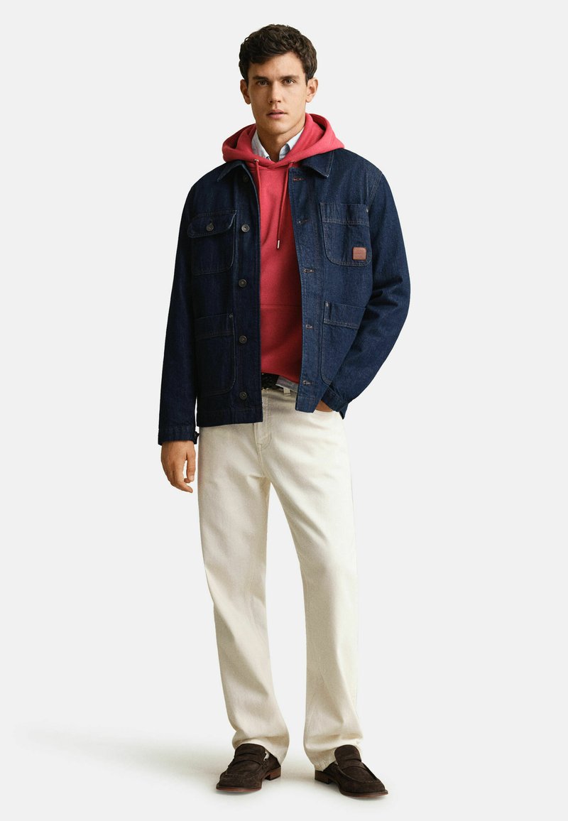 Junger Mann, der einen roten Hoodie unter einer dunkelblauen Jeansjacke, cremefarbene Hosen und dunkelbraune Schuhe trägt, mit einer Hand in der Tasche stehend.