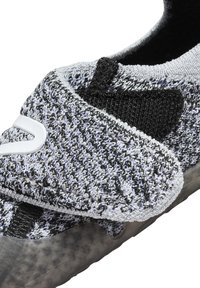Zapato de slip-on en blanco y negro con un upper de punto texturizado, detalles elásticos y una suela de goma. Cuenta con un cierre de Velcro.