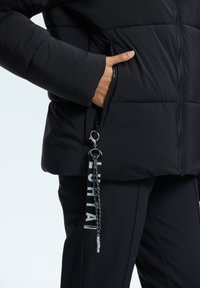 Schwarze Steppjacke mit einer Reißverschlusstasche, ausgestattet mit einem glänzenden silbernen Reißverschluss und einem dekorativen Schlüsselanhänger. Der Stoff hat eine glatte, matte Oberfläche.