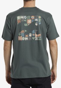 Camiseta de algodón verde oscuro con un patrón de bloques colorido en la parte posterior, con varias formas geométricas y diseños a mano. Manga corta, cuello redondo.
