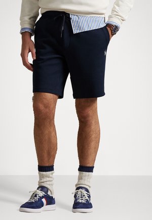 Pantaloni corti in cotone blu navy con vita elastica, cordoncino, tasche laterali e logo, abbinati a sneakers blu e calzini beige.