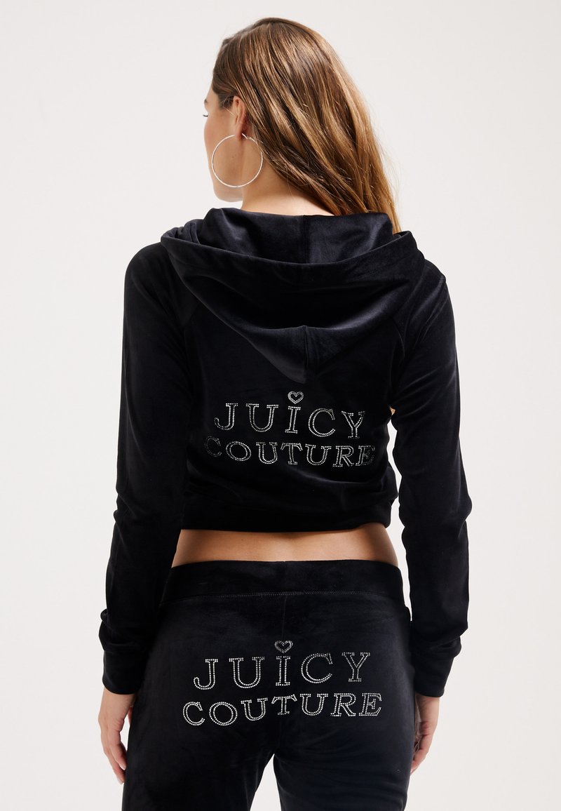 Tuta in velluto nero composta da giacca cropped con cappuccio e pantaloni coordinati. Entrambi i capi riportano "JUICY COUTURE" in strass sul retro.