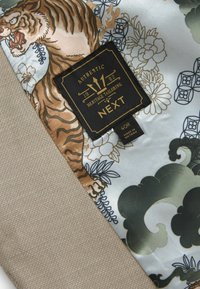 Seidenstoff zeigt einen illustrierten Tiger und florale Muster in gedämpften Farben. Ein strukturierter beiger Akzent und ein schwarzes Etikett mit goldenen Details sind sichtbar.