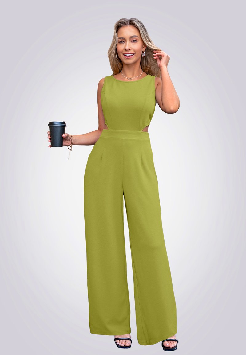 Olijfgroene jumpsuit met mouwloos ontwerp, wijde pijpen en zijuitsparingen. Model houdt een zwarte beker vast, draagt zwarte sandalen en is voorzien van sieraden.