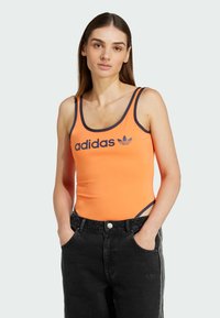Oranje sporttanktop met marineblauwe afwerking en Adidas-logo, gecombineerd met hooggerokken zwarte jeans. Heeft een aansluitend ontwerp en zij-snijdingen.
