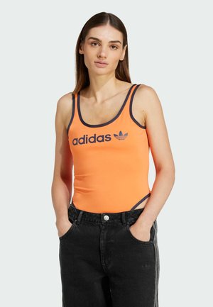 Pomarańczowy sportowy top na ramiączkach z granatowym wykończeniem i logo Adidasa, w połączeniu z czarnymi jeansami z wysokim stanem. Posiada dopasowany krój i wycięcia z boku.