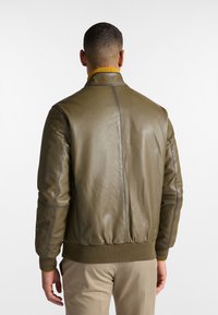 Olivenfarbene Leder-Bomberjacke mit geripptem Saum und Kragen, die gelbe Akzentnähte und eine glatte Textur aufweist.