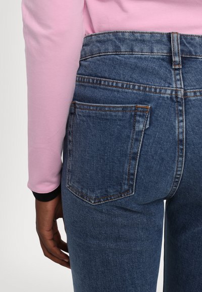 Blue denim jeans with a high waist, defined back pockets, and a faded texture, paired with a pink long-sleeve top featuring black cuffs.